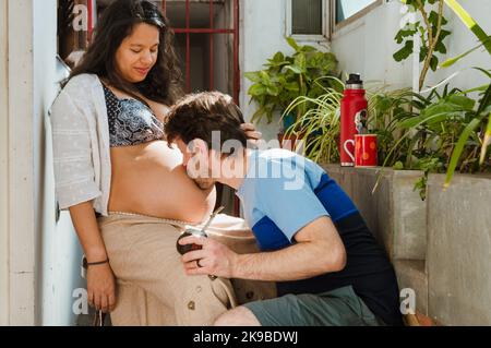 giovane uomo caucasico latino a casa baciando il ventre di sua moglie che è incinta e seduto felicemente guardando come dà l'amore al suo prossimo bambino. Foto Stock