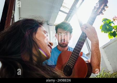 primo piano di un giovane argentino latino con barba, sta guardando e cantando canzoni con la sua chitarra acustica a sua moglie a casa, con t Foto Stock