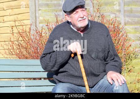 uomo anziano o anziano seduto su una panchina rilassante. Uomo che indossa un cappuccio di stoffa e che tiene un bastone da passeggio. Foto Stock