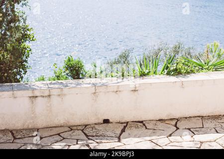 confine bianco in cemento per un sentiero turistico con piante verdi su di esso in un sentiero turistico che conduce lungo l'acqua blu. Foto Stock