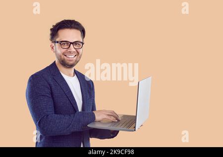 Uomo d'affari felice in tuta e occhiali in piedi su sfondo beige, tenendo il computer portatile e sorridendo Foto Stock
