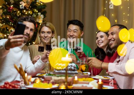 Una compagnia di persone interrazziali che festeggiano insieme il nuovo anno, seduti al tavolo delle feste. L'uomo sta tenendo il telefono, tutti parlano in videochiamata con gli amici, salutano, sorridono. Foto Stock