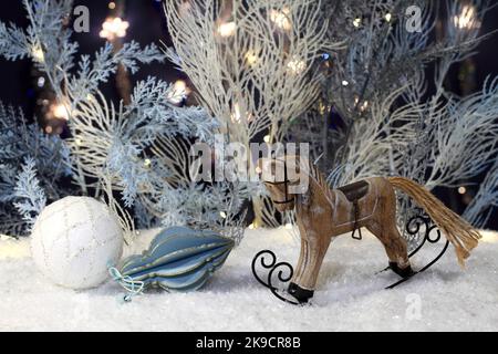 Il cavallo a dondolo giocattolo di legno si trova sulla neve artificiale e accanto a esso si trovano i giocattoli del capodanno e gli alberi artificiali blu e bianchi con le luci festive si trovano behi Foto Stock