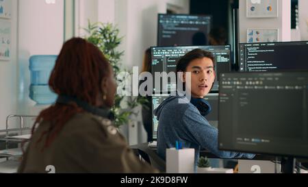 Ingegnere di sistema asiatico che chiede aiuto al codificatore con il codice html, puntando allo script del database sulla finestra del terminale. Team di sviluppatori diversi che lavorano al programma di sviluppo di app nella sala dati. Foto Stock