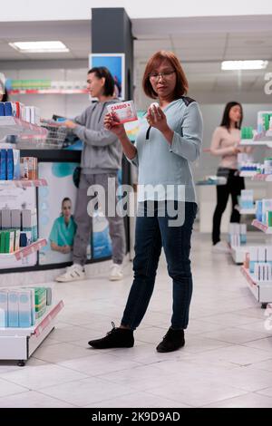 Cliente anziano che guarda le pillole di cardiologia, leggere il foglio illustrativo sulle scatole in farmacia farmaceutica con varie medicine sugli scaffali. Esame di farmaci e pacchetti farmaceutici in farmacia. Foto Stock