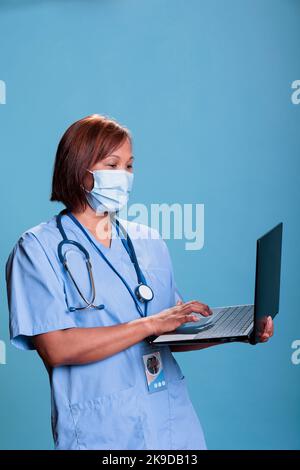 Assistente medico indossare maschera protettiva per prevenire l'infezione con covid19 tenere computer portatile controllo esperienza virus. Infermiere in uniforme blu che lavora alla diagnosi durante la visita di controllo Foto Stock