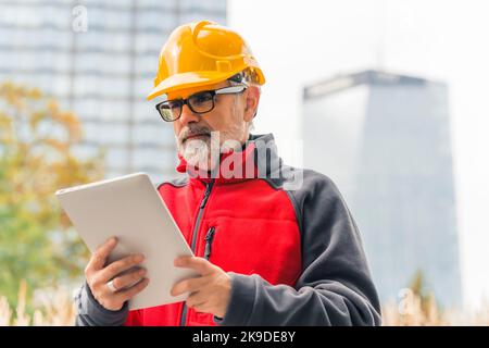 Operaio di costruzione professionale caucasico, bearded, grigio-haired in uniforme che indossa un casco arancione brillante e gli occhiali, in piedi, tenendo una tavoletta usata per lavoro. Foto di alta qualità Foto Stock