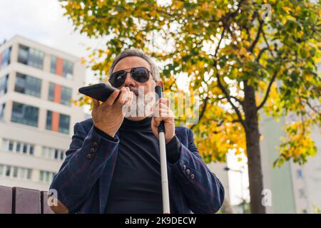 Uomo cieco di mezza età con capelli e barba grigi che indossa occhiali da sole e tuta seduto sul banco che tiene la canna bianca e parla al telefono. Scatto orizzontale all'aperto. Foto di alta qualità Foto Stock