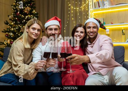 Una compagnia di giovani interrazziali. Sono seduti in un appartamento festoso, con bicchieri di alcool, per celebrare il nuovo anno. Stanno guardando la macchina fotografica, vestita di cappelli rossi di Babbo Natale. Foto Stock