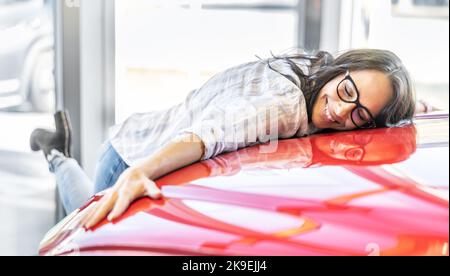 Donna eccitata che abbraccia la sua nuova auto rossa da sogno in una concessionaria di auto. Foto Stock