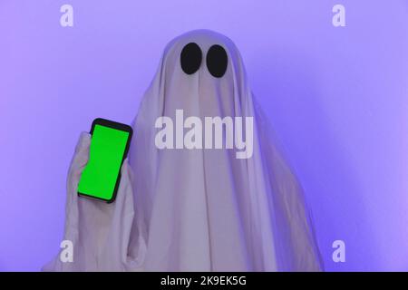 Il fantasma mostra un telefono con uno schermo verde. Telefono con chromakey. Il fantasma fa un ordine da smartphone su Halloween. Spazio libero. Il fantasma si pone con il telefono. Foto Stock