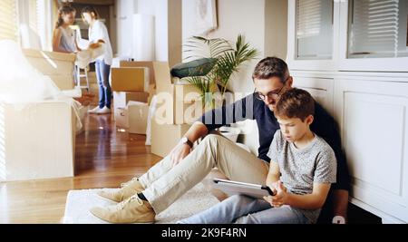 Cercare di connettere e lavorare il wifi. Un padre e suo figlio usano un tablet digitale mentre si trasferiscono nella loro nuova casa con la famiglia. Foto Stock