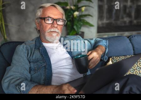 Ritratto di un uomo d'affari anziano con una tazza di caffè e un computer portatile seduti sul divano a casa o in un ufficio moderno Foto Stock
