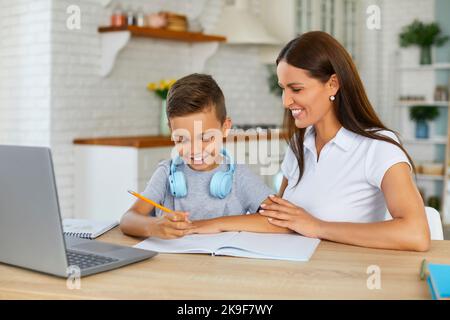 la mamma sorridente sta aiutando il figlio a fare i compiti a casa studiando online utilizzando il notebook in cucina. Foto Stock