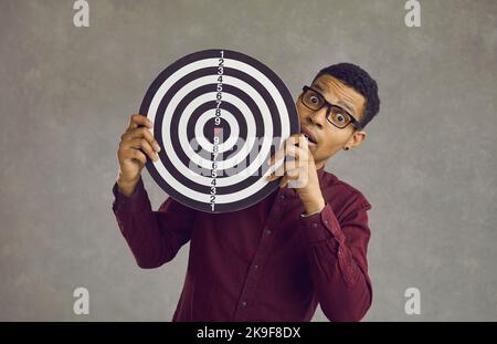 Il giovane afro-americano ha scioccato l'uomo con il dartboard sopra la parete grigia dello studio Foto Stock