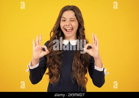 Faccia eccitata. Sorridente ragazza adolescente, giovane adolescente che mostra ok mano segno e winking guardando la macchina fotografica isolato su sfondo giallo studio. Stupito Foto Stock