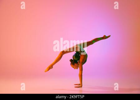 Giovane ragazza teen flessibile ginnastica ritmica in movimento, azione isolata su sfondo rosa in luce al neon. Sport, bellezza, competizione, flessibilità Foto Stock