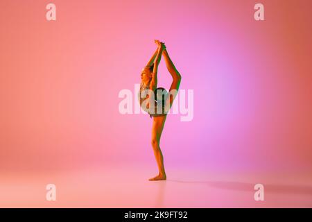 Giovane ragazza teen flessibile ginnastica ritmica in movimento, azione isolata su sfondo rosa in luce al neon. Sport, bellezza, competizione, flessibilità Foto Stock