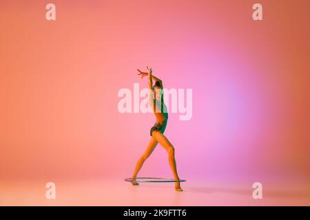 Grazia. Giovane ragazza teen flessibile ginnastica ritmica in movimento, azione isolata su sfondo rosa in luce al neon. Sport, bellezza, competizione Foto Stock