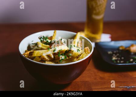 Ciotola di patatine fritte gourmet all'interno di un ristorante con luce soffusa e birra sullo sfondo. Elegante contorno o antipasto con salsa e erba cipollina e parmesa Foto Stock