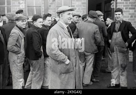 1938, storici lavoratori industriali che fanno la fila fuori dal loro posto di lavoro per la retribuzione settimanale, con caposquadra e cappello piatto, Treforest Industrial Estate, Rhondda, Galles del Sud, Regno Unito. Le origini della tenuta risiedono nel South Wales & Monmouthshire Trading EstaIl patrimonio ha origine nel South Wales & Monmouthshire Trading Estates Ltd, una società governativa senza scopo di lucro fondata per creare e diversificare l'occupazione nella zona, lontano dalle tradizionali industrie del carbone e dell'acciaio che erano in declino a lungo termine. A metà degli anni '1940s, circa 16.000 persone erano occupate nella proprietà commerciale. Foto Stock