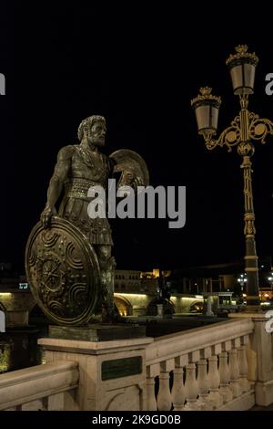 Statue che decorano il Ponte delle civiltà, di fronte al Museo Archeologico di Skopje, Macedonia settentrionale. Di notte con lampioni che illuminano il ponte. Foto Stock