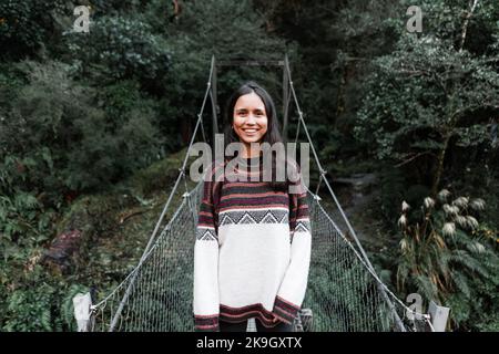 calma e rilassata brunette giovane donna caucasica sorridente in piedi su ponte di metallo filo che attraversa il fiume in mezzo alla giungla e la foresta, specchio lago, nuovo Foto Stock