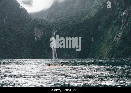 due persone in kayak e fare sport nelle acque calme del lago vicino alle imponenti montagne circondate da alberi e una bella cascata Foto Stock