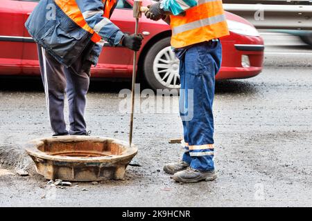 I lavoratori della strada in tute arancioni stanno riparando un vecchio tombino delle fognature sulla carreggiata contro uno sfondo sfocato di un'auto di passaggio. Spazio di copia. Foto Stock