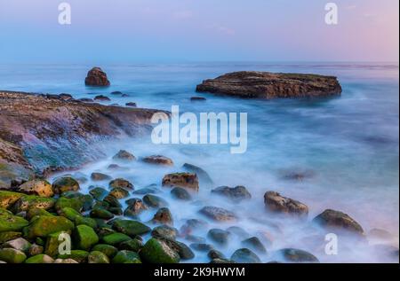 Una tranquilla scena costiera al tramonto caratterizzata da una collezione di rocce aspre ricoperte di muschio, immerse nella calda luce dorata in contrasto con l'acqua dolce da sogno. Foto Stock