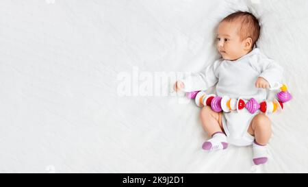 La bambina tiene il giocattolo colorato del sonaglino. Vista dall'alto del ragazzino sdraiato su lino bianco stropicciato con il suo primo giocattolo. Neonato a letto. Banner con spazio di copia. Foto Stock