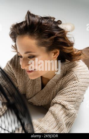 donna brunetta sorridente in cardigan lavorato a maglia sdraiata vicino al ventilatore elettrico soffiato su sfondo grigio Foto Stock