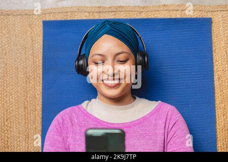 Felice donna biraciale indossando hijab, stendendo su tappetino yoga e utilizzando smartphone in soggiorno Foto Stock