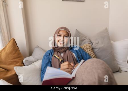 Ritratto di felice donna biraciale indossando hijab, seduto sul divano e prendendo appunti Foto Stock