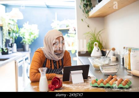 Felice donna biraciale che indossa hijab, in piedi in cucina, utilizzando tablet e cottura Foto Stock