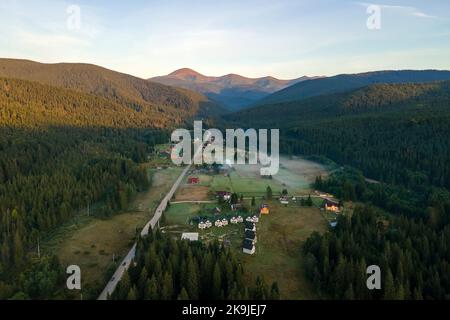 Vista aerea di mattina di nebbia luminosa sopra le piccole case rurali tra cime scure con alberi di foresta di montagna all'alba di autunno. Bellissimo scenario di selvaggio Foto Stock