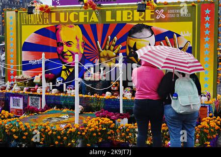 Città del Messico, Messico. 28th Ott 2022. Due persone si nascondono sotto e sotto l'ombrello durante una tempesta pomeridiana mentre si guarda un Ofrenda o altare in onore del wrestling messicano durante la Megaofrenda di Zocalo festival per celebrare l'inizio della Giornata dei morti a Plaza de la Constitucion, 28 ottobre 2022 a Città del Messico, Messico. Credit: Richard Ellis/Richard Ellis/Alamy Live News Foto Stock