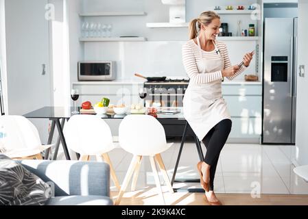 Ricerca di idee per la cena. Una donna matura che usa il suo tablet digitale mentre indossa il grembiule in cucina. Foto Stock