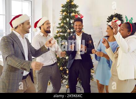 Amici multirazziali felici che celebrano il Natale parlando con gli scintillatori nelle mani vicino all'albero di Natale. Foto Stock