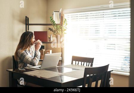 Il caffè, la contemplazione e la connettività sono le modalità con cui inizia la sua mattinata. Una giovane donna beve un caffè e usa un computer portatile la mattina a casa. Foto Stock