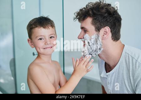 Im preparazione del papà per una rasatura pulita. Ritratto di un ragazzino che si asciuga la crema sul viso dei padri nel bagno di casa. Foto Stock