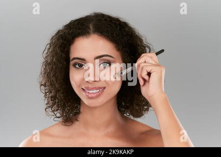 Per una faccia con tono uniforme, applicare correttamente la fondazione. Studio shot di una bella giovane donna che applica fondazione con un pennello da make up contro un grigio Foto Stock