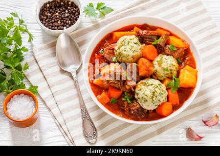 Stufato di manzo con gnocchi e verdure in ricco pomodoro e sugo a base di brodo in ciotola bianca su tavolo di legno, vista orizzontale dall'alto, piatto, libero s Foto Stock