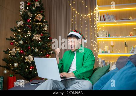Uomo asiatico che legge buone notizie sul notebook a casa per Natale, uomo d'affari che lavora a distanza durante le vacanze di Capodanno seduto sul divano in soggiorno, celebrando il successo della vittoria. Foto Stock