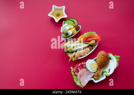 Piatto in forma di albero di Natale con cibo proteico - carne, pesce, formaggi, noci, ecc sfondo rosso. Il concetto di dieta del keto tratta per il holi Foto Stock