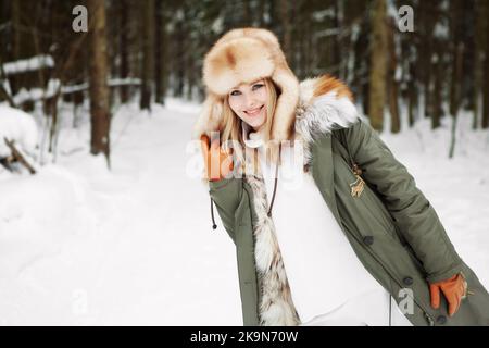 Ritratto di giovane donna in inverno pino nevoso sfondo foresta, indossando pelliccia parka cappotto, orecchie flaps cappello e guanti in pelle brillante, attività sana. Bionda Foto Stock