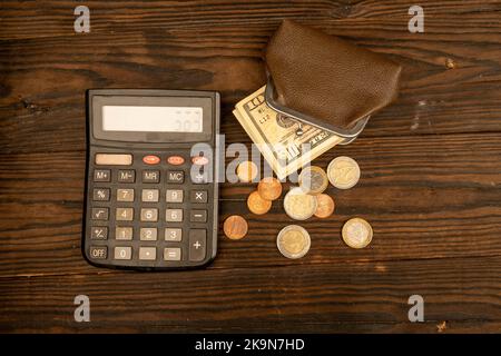 Banconote in dollari, monete in euro, un portafoglio in pelle marrone vintage e una calcolatrice su una superficie di legno. Primo piano, messa a fuoco selettiva. Foto Stock