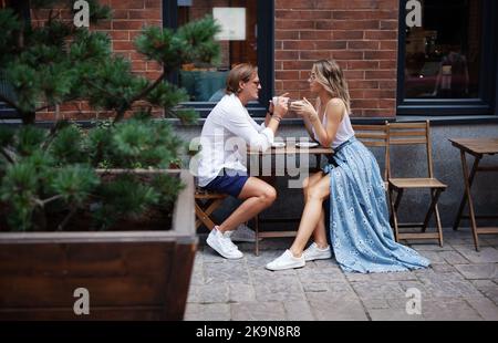 Giovane coppia alla moda che parla al Coffee Shop in un appuntamento. Coppia amorevole che ha tazza di caffè al ristorante di strada. Uomo e donna alla moda portr a lunghezza intera Foto Stock