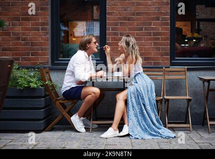 Bella giovane coppia alla moda seduta in un bar e bere caffè fuori casa. Colore naturale. Donna elegante che si nutre con il cucchiaio uomo i Foto Stock