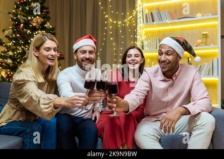 Giovani allegri gruppi di persone, gli amici celebrano il conto alla rovescia di Capodanno a casa appicchiano i bicchieri da vino mentre si siedono sul divano a casa. Si augurano un felice buon Natale e un abbraccio insieme divertendosi Foto Stock